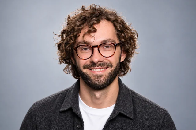 Professional headshot of a man with round glasses and a beard wearing charcoal layers.